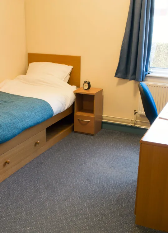 A spacious bedroom. The floor is blue carpeted and a neatly made single bed is visible against one wall. On the other side of the room is a wooden desk with office chair and a large window.