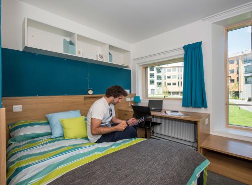 A student sits using a tablet on a large, brightly coloured bed in a modern bedroom. Modern buildings and a grassy courtyard can be seen through the windows.