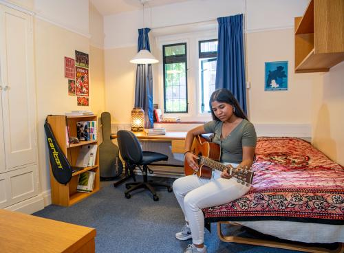 A student plays a guitar while sitting on a bed in a bedroom. In the background, shelves and a desk with books can be seen.