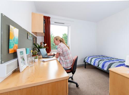 A spacious, bright student bedroom showing a single bed, desk and open window. A student sits at the desk, working on a laptop.