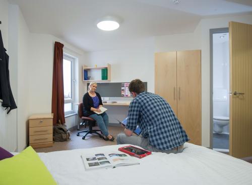 Two students sat talking in a large spacious bedroom. A double bed, desk and open door to a bathroom can be seen.