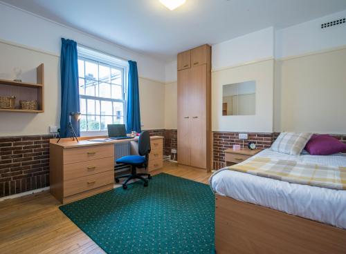 A spacious student bedroom with double bed, green rug on a wooden floor and large desk. Light streams in through a large window.