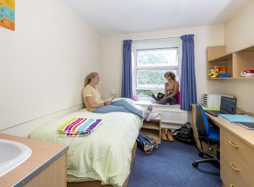 Two students chatting in a bedroom. One sits browsing with her tablet on a single bed while the other looks up from their book, sat on the windowsill.