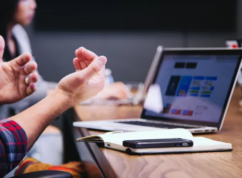 Laptop on a table with notepad and phone, user's hands in foreground 