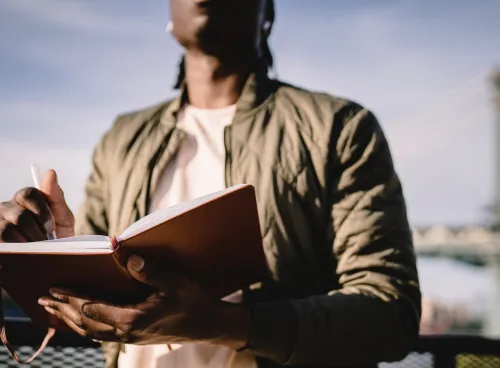 A Black man writes on a notepad as he stands outside, a blue sky and a bridge behind 