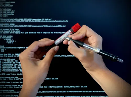 Close-up of hands labelling a patient blood sample for molecular analysis. In the background a computer screen shows programme code to process sequencing data. 