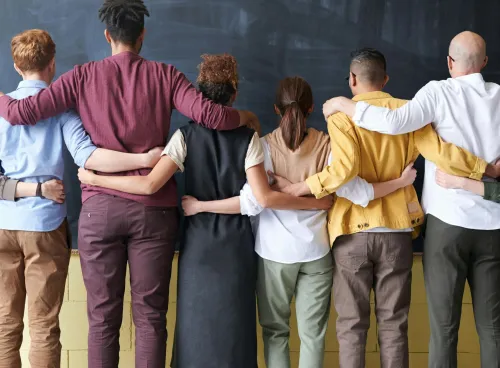 A diverse group of nine people stand in a row with their arms around each other's shoulders, facing a blackboard in a classroom, dressed in an array of casual to smart-casual attire.