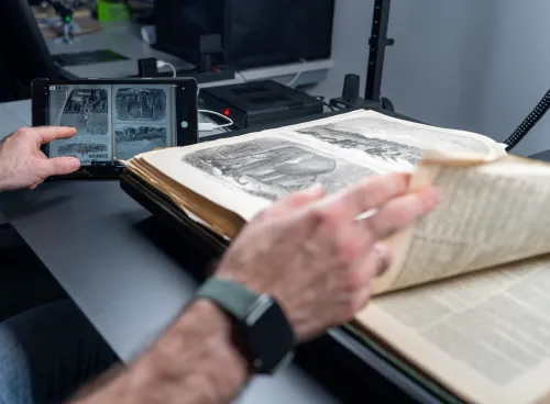 A close-up of a person's hands working with an open historical book on a specialized scanning table. One hand is turning a delicate page with illustrations, while the other holds a tablet displaying a digital scan of the same page. The workspace includes modern scanning equipment and cables in the background, blending traditional and digital technologies.