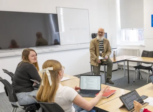 Alt text: A teacher sits on a desk holding papers while three students face them, one using a laptop. A whiteboard behind the teacher displays the phrase "POINT OF VIEW."