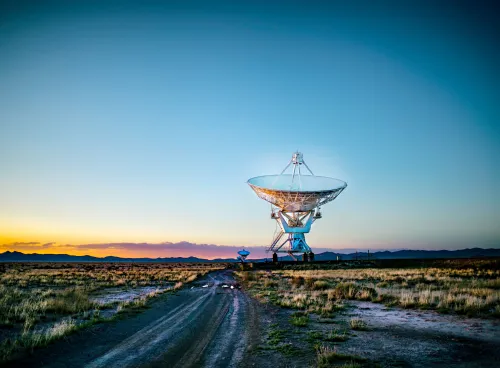 A towering satellite dish positioned in a wide, grassy field, silhouetted against a vivid orange sunset sky, with a winding dirt road leading toward it.