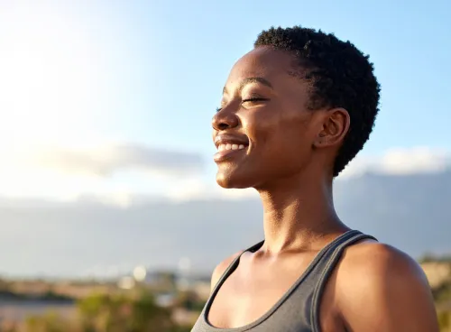 Person with short hair smiling with eyes closed against a bright sky background.