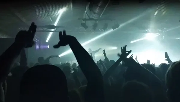 Silhouette of large crowd with their hands in the air, against bright lights of nightclub.