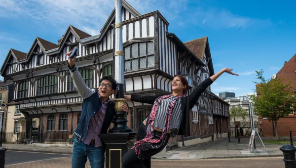 Two students take a selfie outside a large classic tudor house.