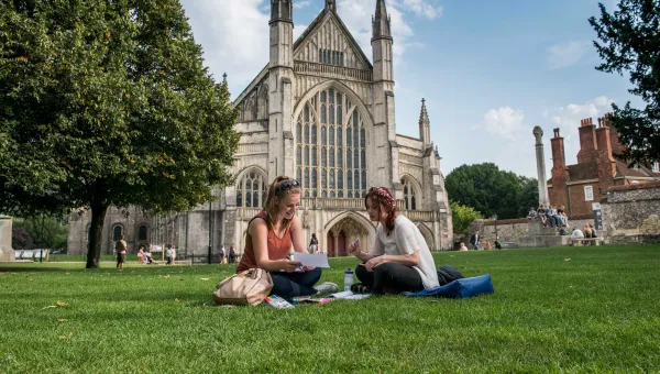 Two students sit talking on the grass outside a large, ornate cathedral.