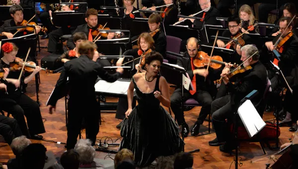 Female opera singer performing, surrounded by members of an orchestra.