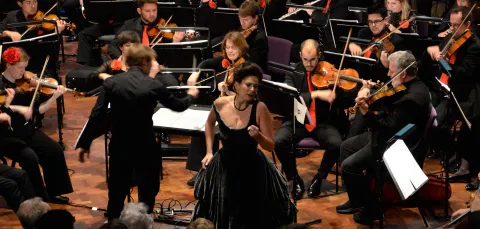 Female opera singer performing, surrounded by members of an orchestra.