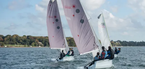 Three small sailing boats, each with a crew of three, on open water beneath blue skies. An area of trees is visible in the background.