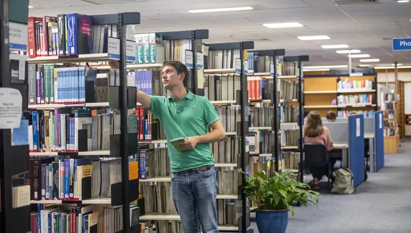 A student selects a book from a shelf while other students work at desks in the background