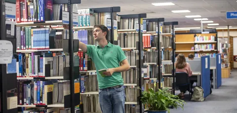 A student selects a book from a shelf while other students work at desks in the background