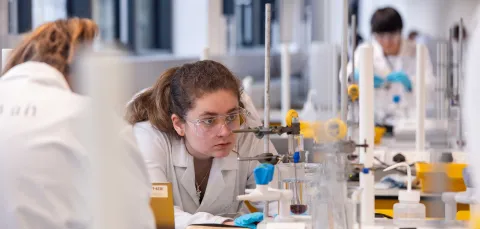 A student in a lab coat crouches down to closely monitor a chemistry experiment being conducted in a large piece of glassware.