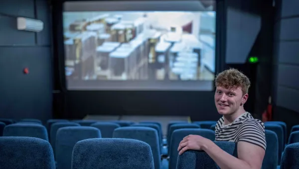 A student leaning over the back of his seat in a cinema as a film plays in the background, smiling at the camera.