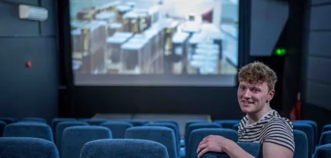 A student leaning over the back of his seat in a cinema as a film plays in the background, smiling at the camera.