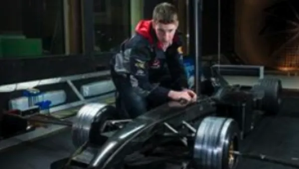 A male student kneeling beside a half-scale model of a Formula 1 car, adjusting various controls.