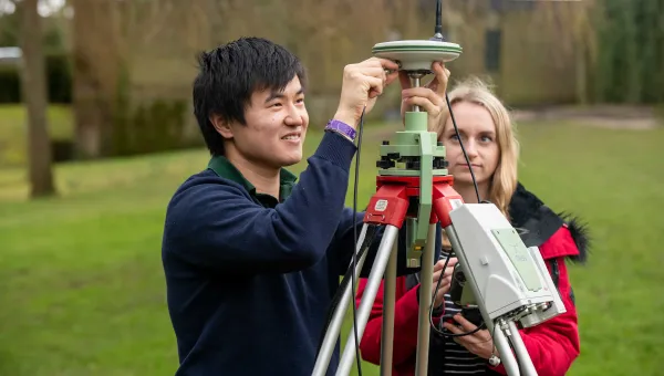 Two students conduct an experiment outdoors using a large piece of survey equipment on a tripod.