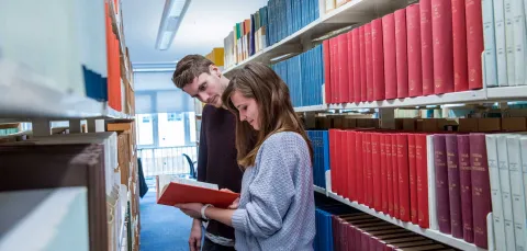 Two students in a narrow library aisle, surrounded by shelves of books on both sides. They are reading a book pulled from one of the shelves.