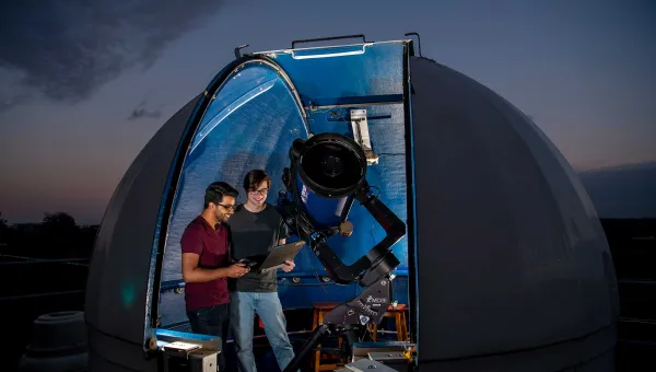 A small, domed rooftop observatory opens onto a starry night sky. Inside the observatory, two students use a telescope and a laptop to collect data.