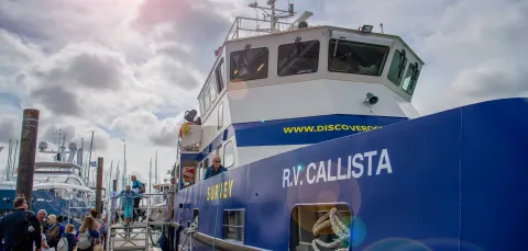 An industrial looking boat emblazoned with the name RV Callista, moored beside a wooden dock, beneath a sunny sky.