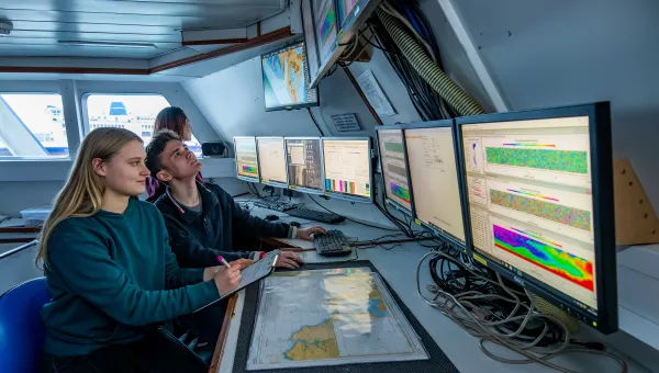Two students sit at a desk inside a room on a boat. They observe a bank of computer screens displaying various data and information.