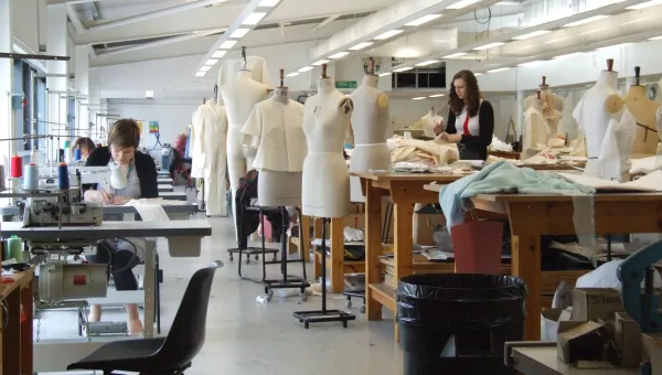A wide shot of the fashion studio, full of students hard at work. To the right, a row of large workbenches are mixed in with over a dozen mannequins wearing various designs. To the left, a row of sewing machines.