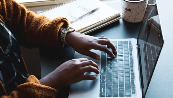 Fingers typing on a laptop with notepad, pen and mug in the background