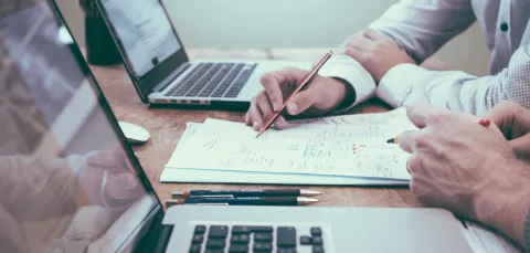 Close up view of two people working at a desk with laptops, paper and pens