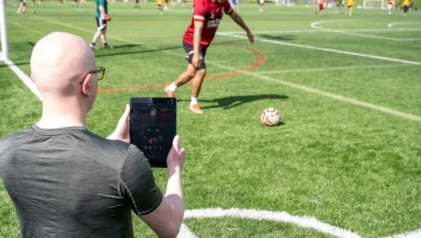 Dr Devon Lewis standing by a football pitch as a player kicks the ball. Devon is holding an iPad that shows live data streamed from a wearable sensor.