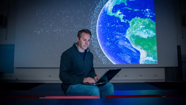 Professor Hugh Lewis sitting on the front bench of a lecture theatre as he consults his laptop. A graphic of satellites orbiting Earth is projected on the screen behind him.