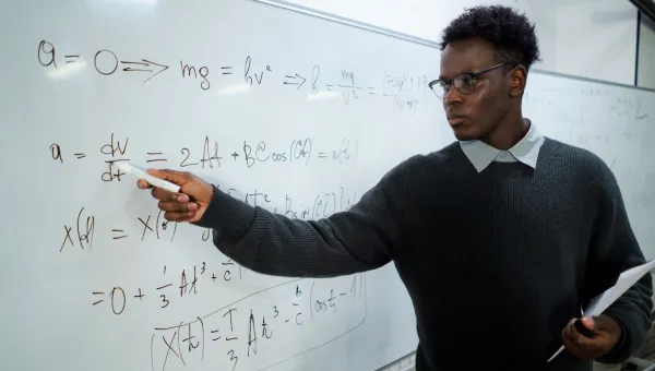 A maths teacher writes on a white board and looks intently at the equations. We imagine he is in front of a classroom of pupils.