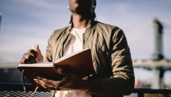 A young Black man, whose face is unseen, writes in a notepad. He has a blue sky and a bridge that is out of focus behind him. The photo focuses on his hand with the pen.