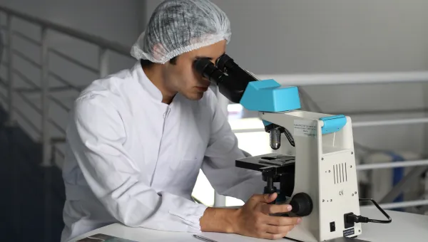 A researcher in a white lab coat looks through a microscope.