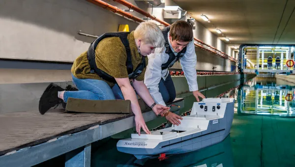 A student crouches down to place a model boat into the water of the university towing tank, a researcher at her side