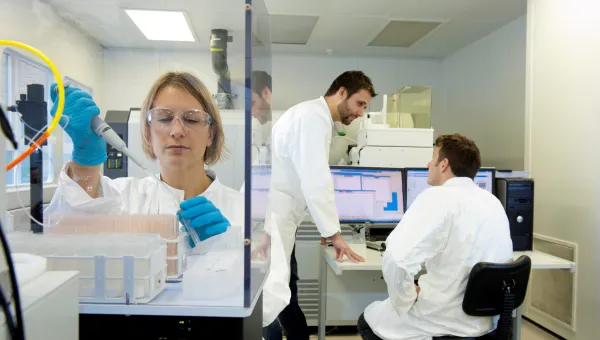 Researcher pipetting in a lab while two researchers talk in the background