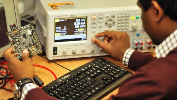 A man in a brown jumper examines a circuit board at a desk