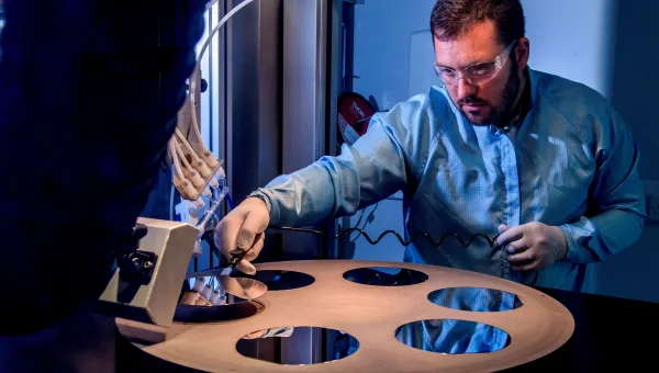 A bearded young scientist in protective overalls and glasses works with a flame hydrolysis deposition (FHD) system