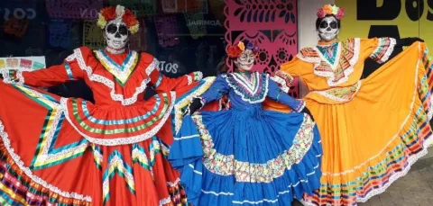 Three dancers wearing brightly coloured Mexican dresses and headgear pose for a photograph