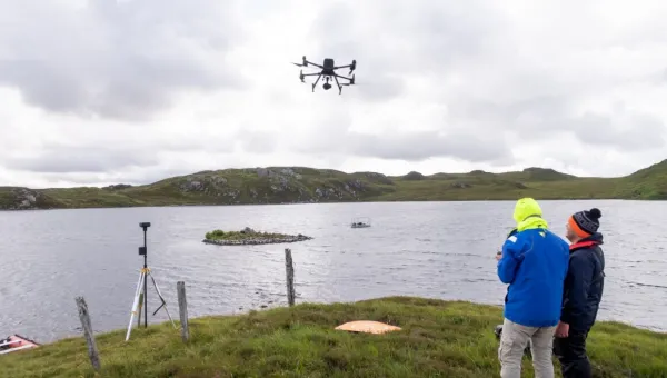 Two archaeologists guide a drone into the air over a grey stretch of water between grassy, low-lying islands. They are well wrapped up against the cold. 