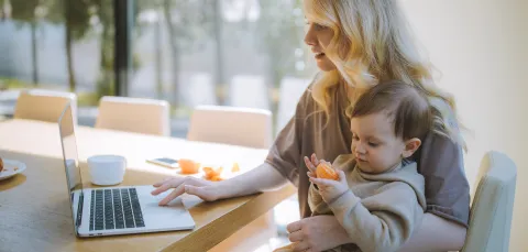 A woman sitting at a table using a laptop with one hand and a child sitting on her knee eating an orange