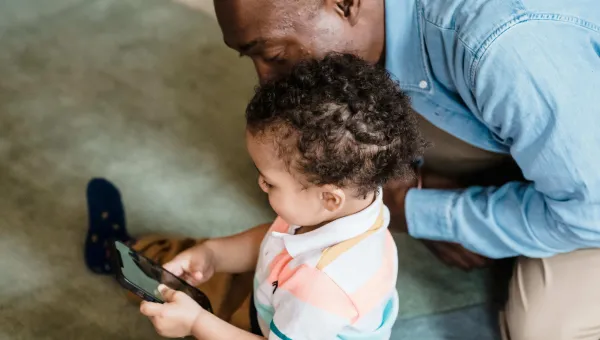 Young child playing on a phone as an adult watches them over their shoulder looks 