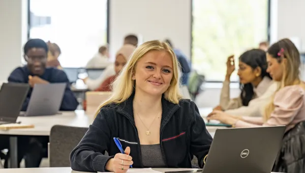 A student smiling while sat at a desk in a classroom. They are holding a pen and they have a laptop and notepad on the table. There are other students with laptops sat at desks in the background.