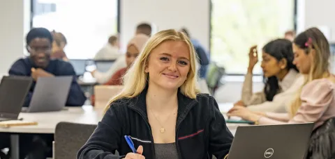 A student smiling while sat at a desk in a classroom. They are holding a pen and they have a laptop and notepad on the table. There are other students with laptops sat at desks in the background.